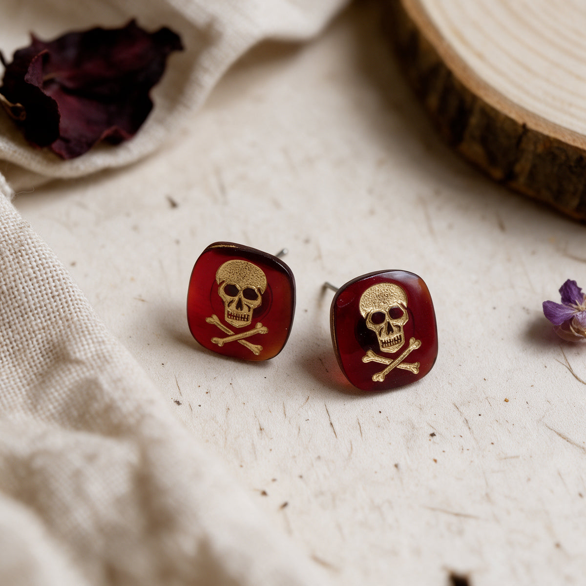 A pair of square stud earrings with a glossy dark red resin background and a raised gold skull and crossbones motif, displayed on a textured light beige surface with dried flowers and fabric.
