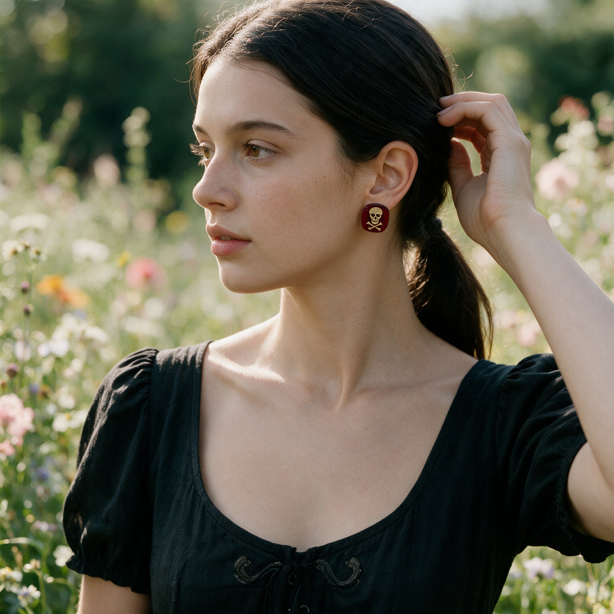 A woman in a black dress stands in a sunlit field of flowers, wearing a small gold skull earring with a red circular background.
