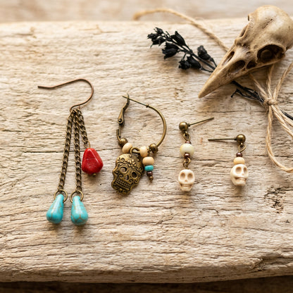 Four bohemian-style dangle earrings displayed on a rustic weathered wood surface alongside decorative props, including a bird skull, dried black flowers, and twine. The earrings feature antique bronze metal, turquoise blue beads, a red stone, skull charms, and cream beads.