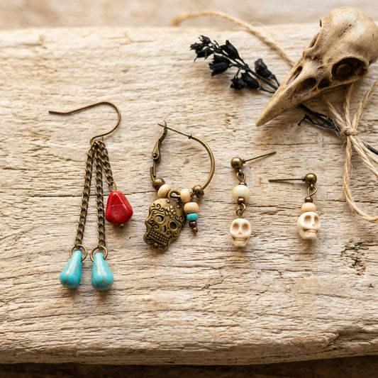 Four bohemian-style dangle earrings displayed on a rustic weathered wood surface alongside decorative props, including a bird skull, dried black flowers, and twine. The earrings feature antique bronze metal, turquoise blue beads, a red stone, skull charms, and cream beads.