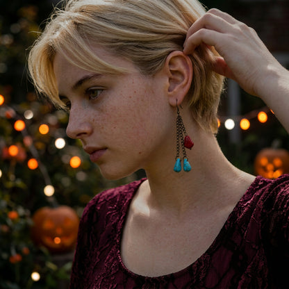 A close-up lifestyle shot of a woman with short blonde hair wearing a long dangle earring. The earring features a dark metal chain, a small red heart charm, and two dangling turquoise blue beads.