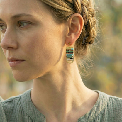Close-up of a woman wearing a gold earring with a scenic design, outdoors.