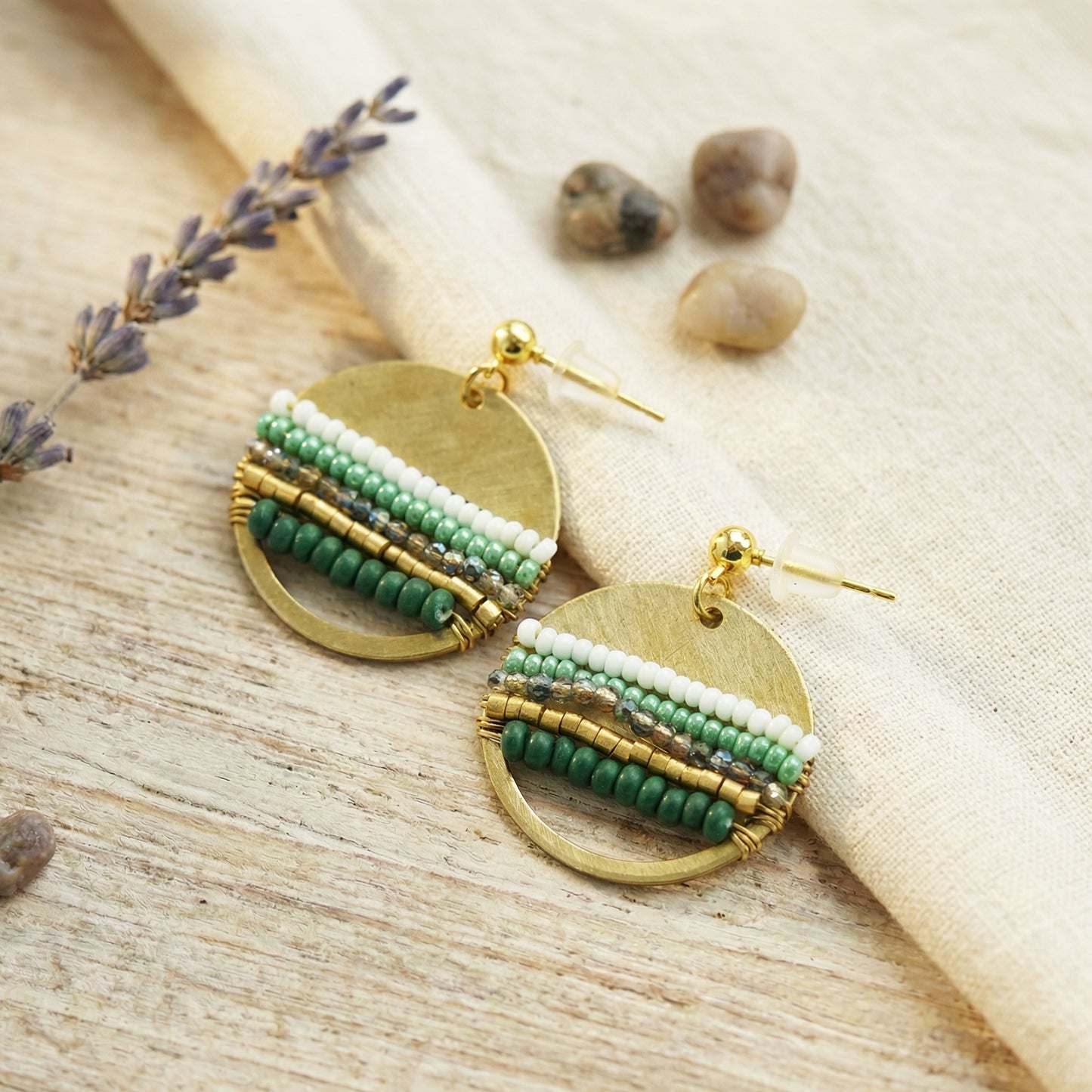 Gold and green beaded earrings on a natural wooden surface with lavender and stones.