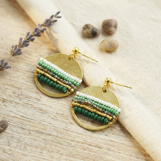Gold and green beaded earrings on a natural wooden surface with lavender and stones.