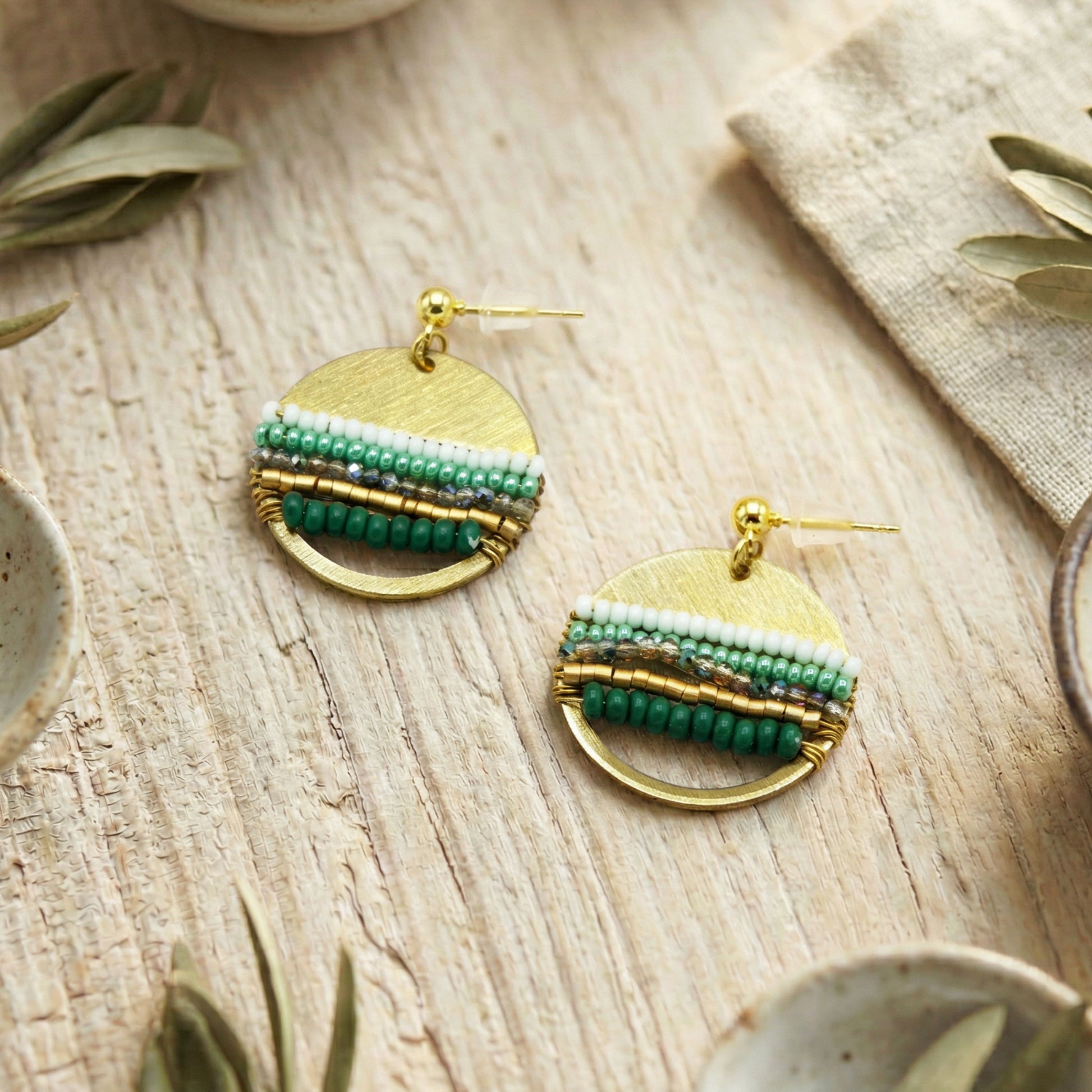 Gold and green beaded earrings on a wooden surface with natural elements.