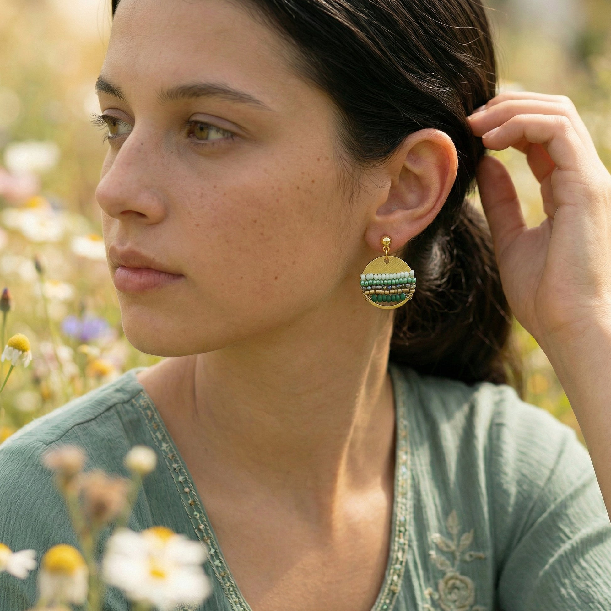 Woman wearing green and gold earrings in a natural setting with flowers.