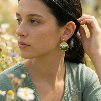 Woman wearing green and gold earrings in a natural setting with flowers.