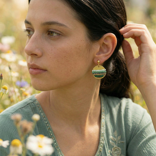 Woman wearing green and gold earrings in a natural setting with flowers.