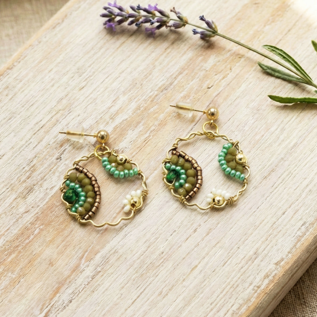 Pair of gold and green beaded earrings on a wooden surface with lavender flowers.