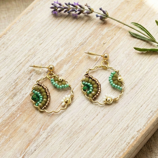 Pair of gold and green beaded earrings on a wooden surface with lavender flowers.