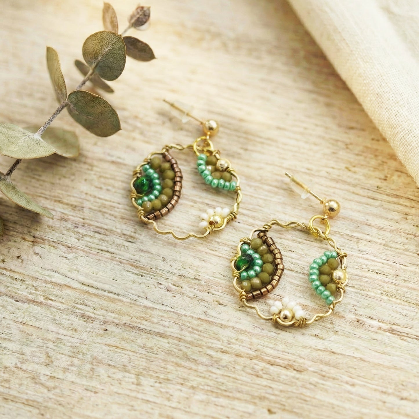 Pair of gold and green beaded earrings on a wooden surface with eucalyptus leaves.