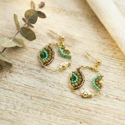 Pair of gold and green beaded earrings on a wooden surface with eucalyptus leaves.