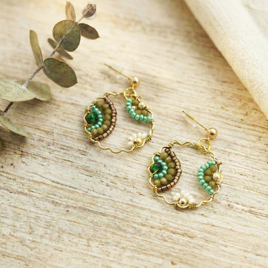 Pair of gold and green beaded earrings on a wooden surface with eucalyptus leaves.