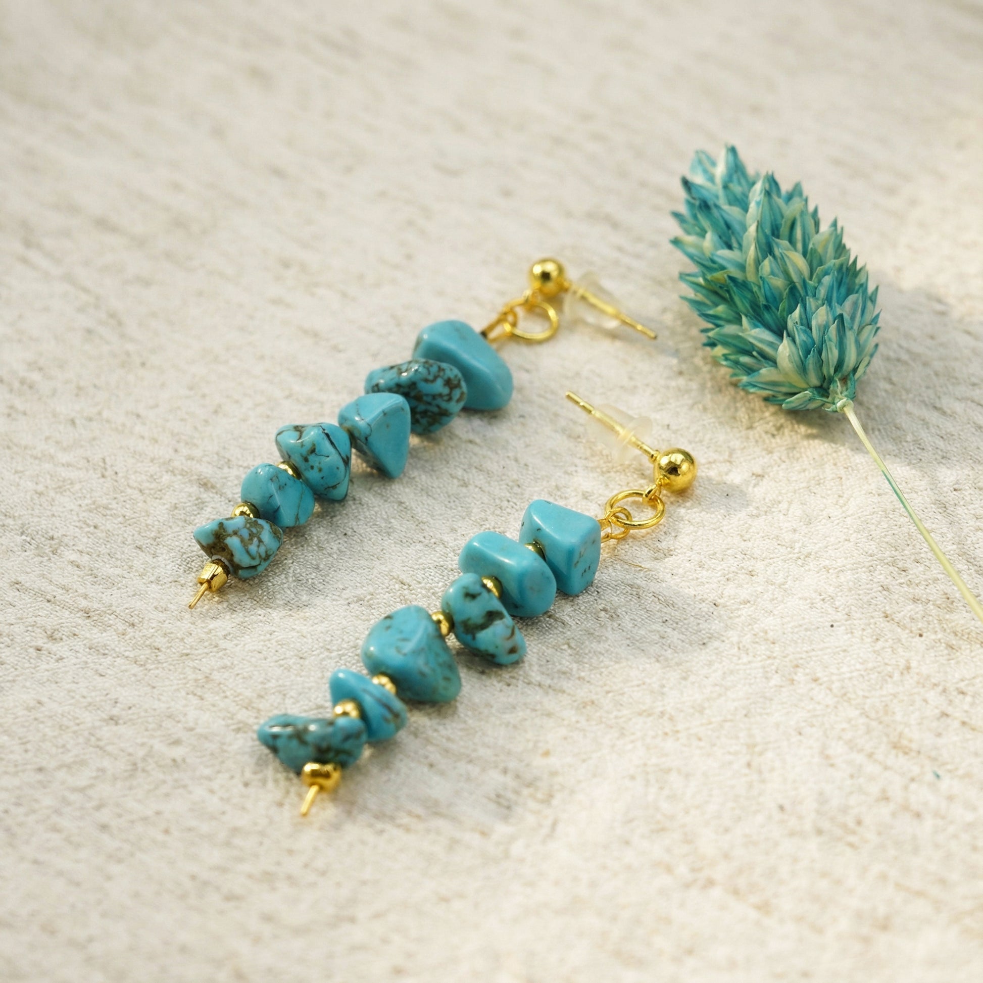 Turquoise stone earrings with gold accents on a textured beige surface with a dried plant.