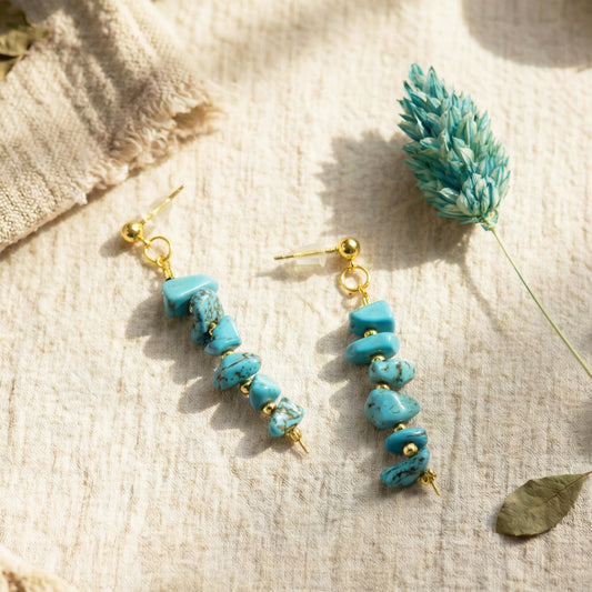 Turquoise stone earrings with gold accents on a textured beige surface with a dried plant.