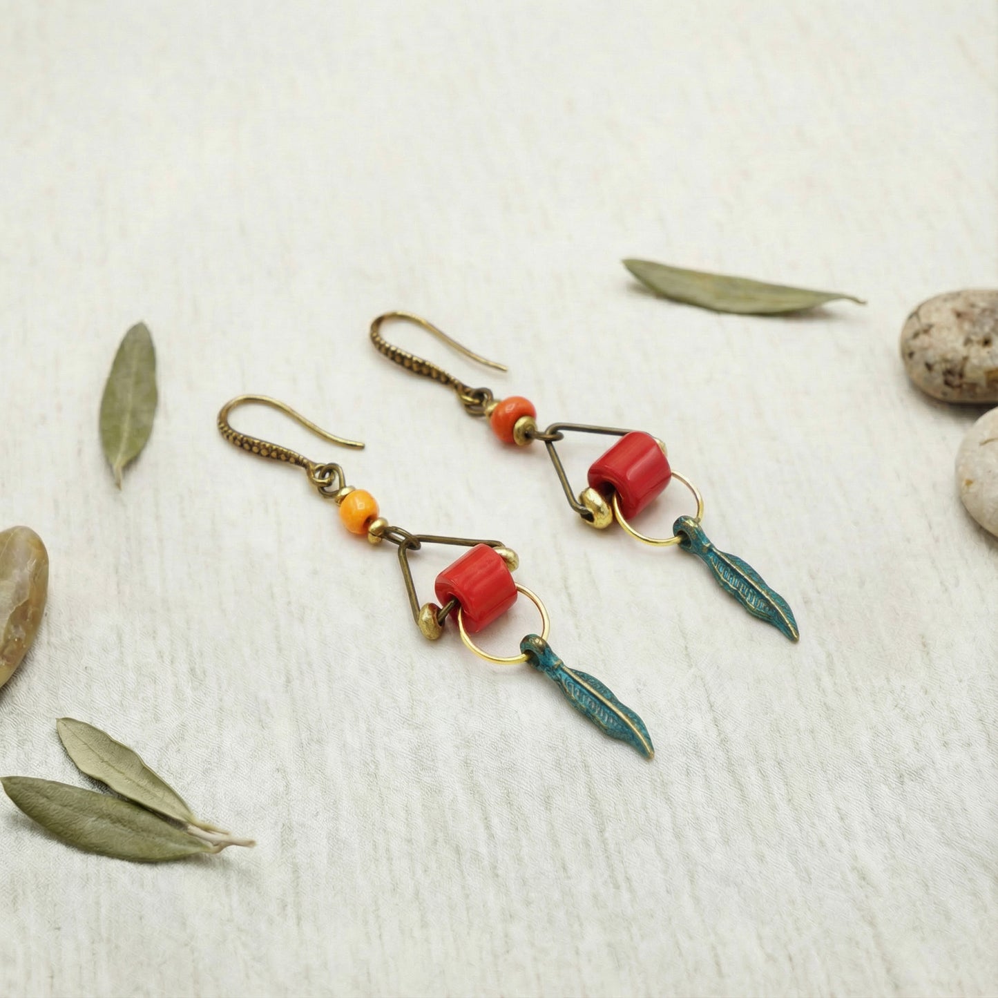 Pair of earrings with red and green accents on a textured surface with leaves and stones.