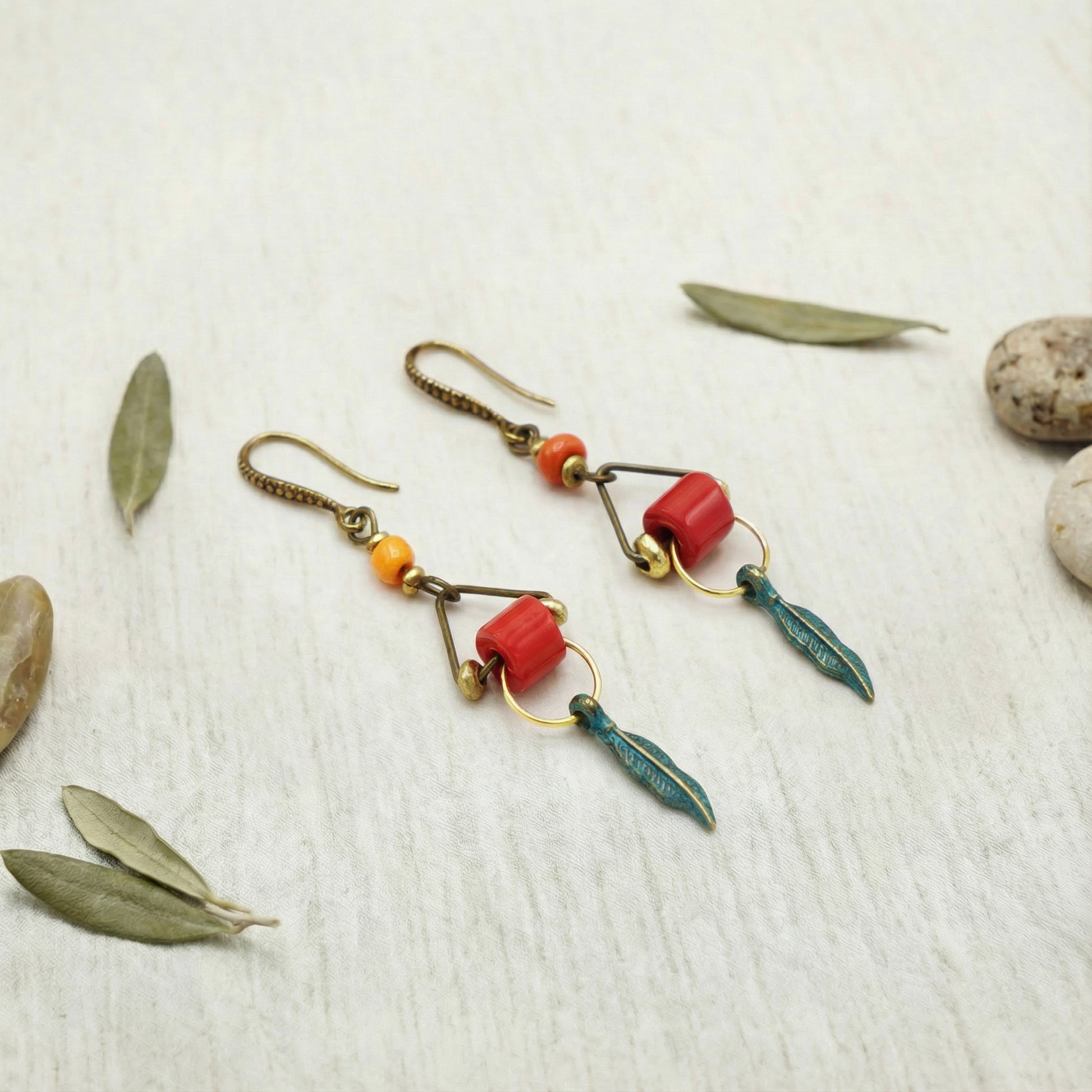 Pair of earrings with red and green accents on a textured surface with leaves and stones.