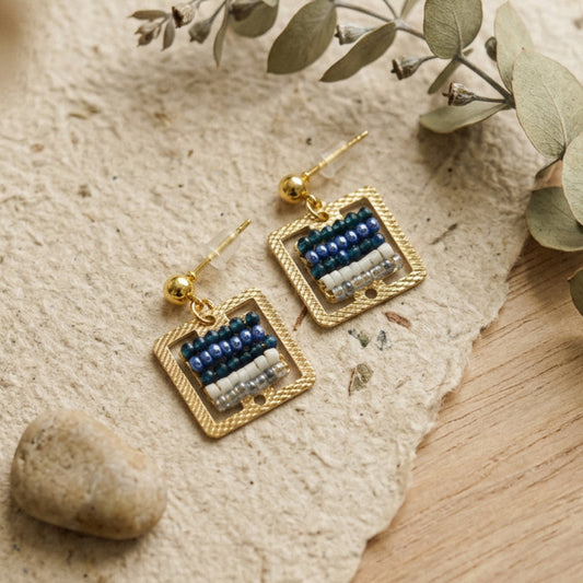 A pair of gold-tone square dangle handwoven earrings with blue and white beaded stripes, displayed on a textured beige stone surface with dried green leaves and a small stone.