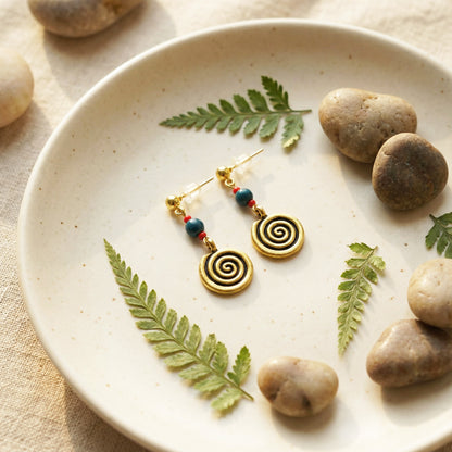 Gold spiral earrings with colorful beads on a plate with stones and leaves