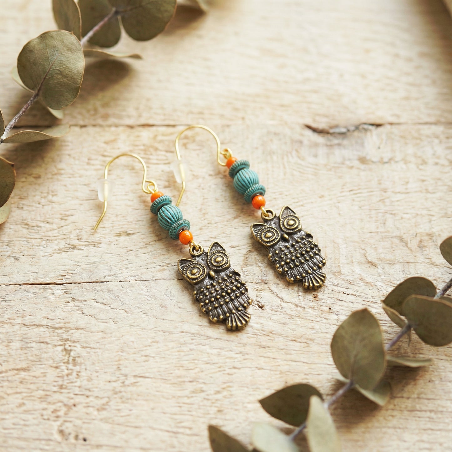 Owl-shaped earrings with turquoise and orange beads on a wooden surface with eucalyptus leaves.