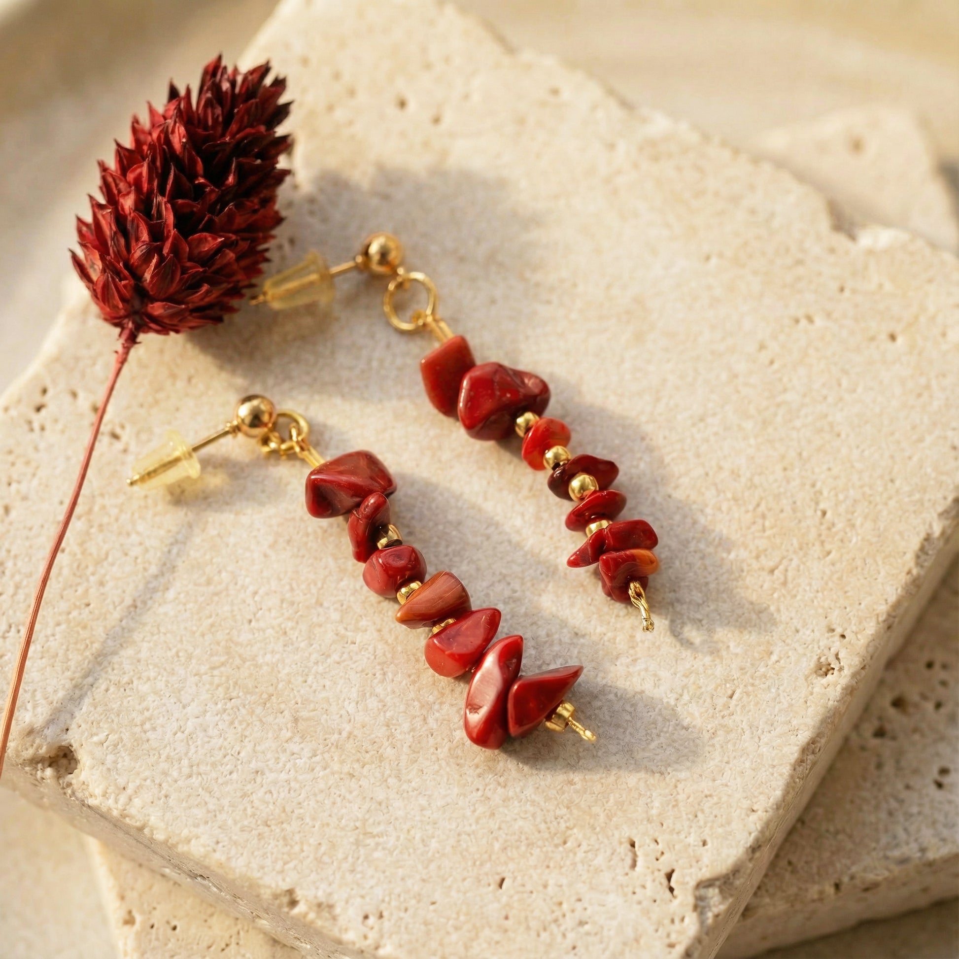 Red stone earrings on a textured beige surface with a dried red flower.